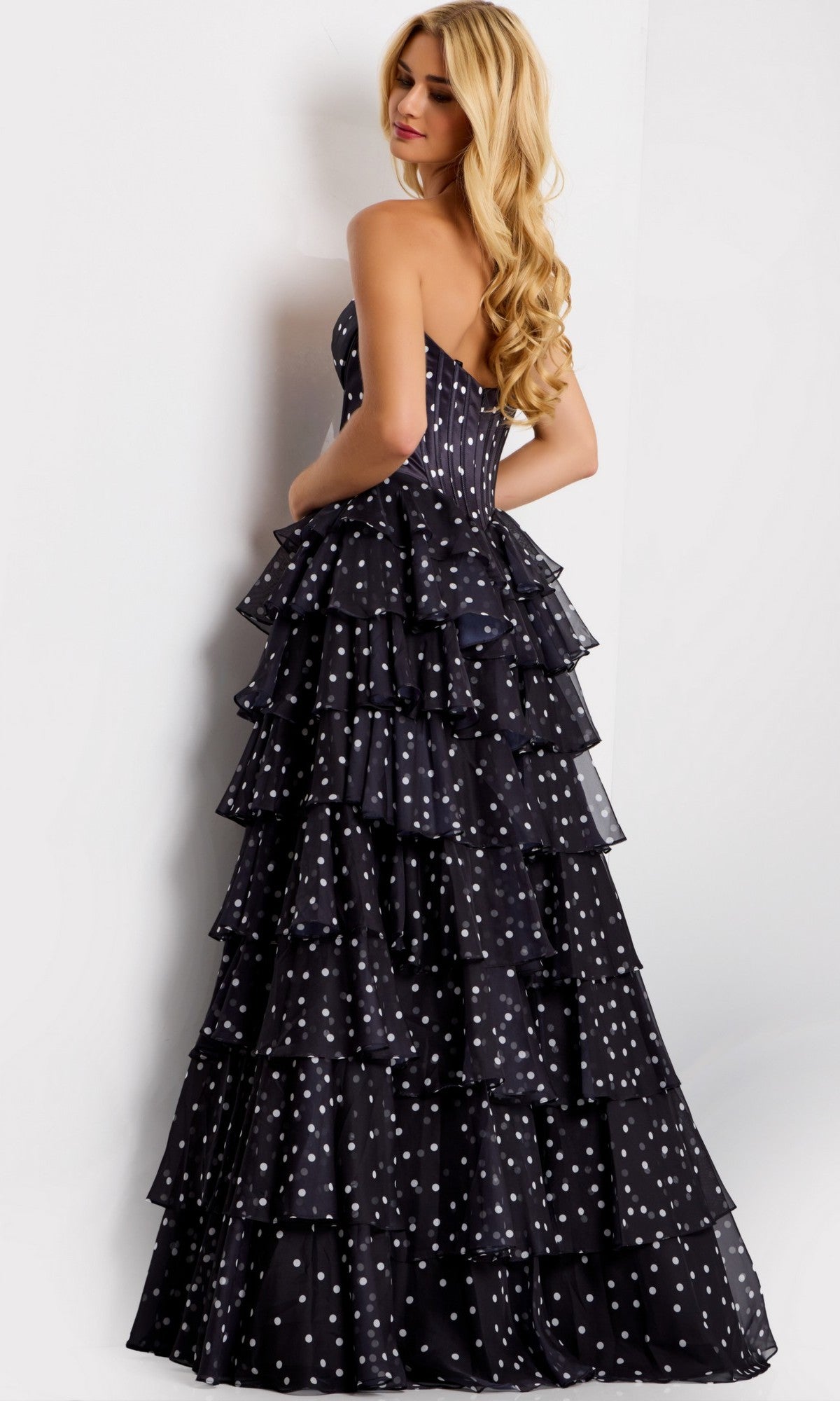 A woman wearing the Long Prom Dress: Jovani 49087—a strapless, floor-length black gown with white polka dots and tiered ruffles—stands against a plain white background.
